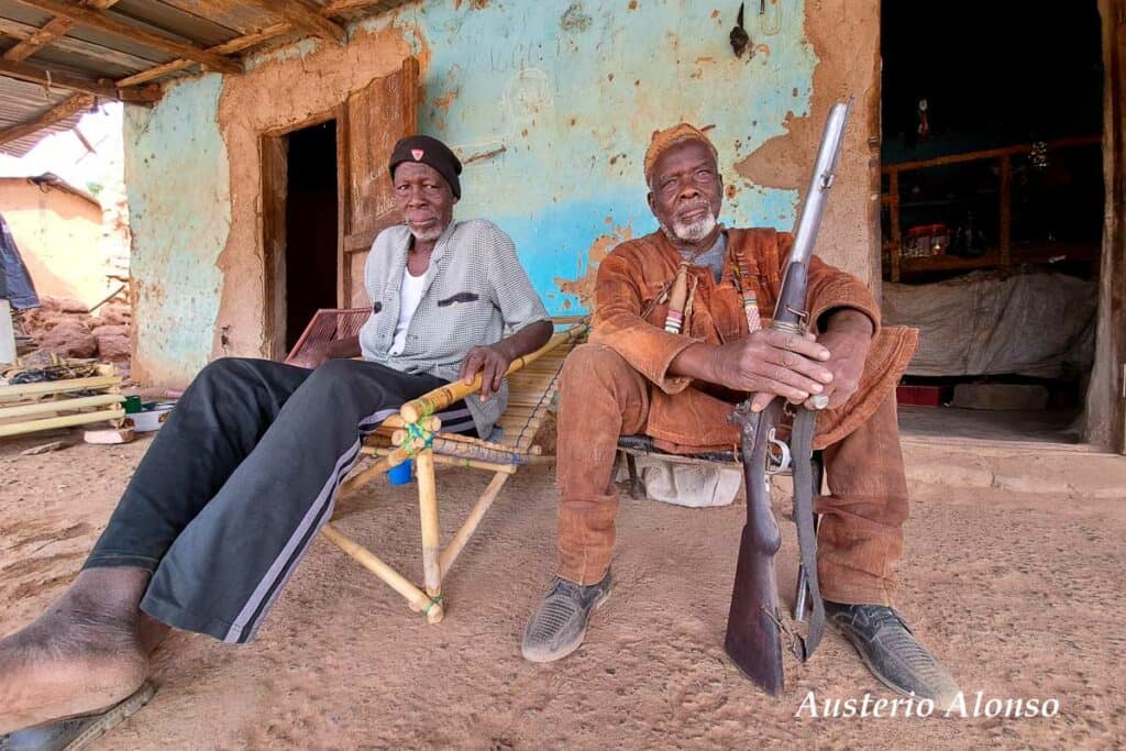two dozo men sitting in hammocks, one of them with a rifle